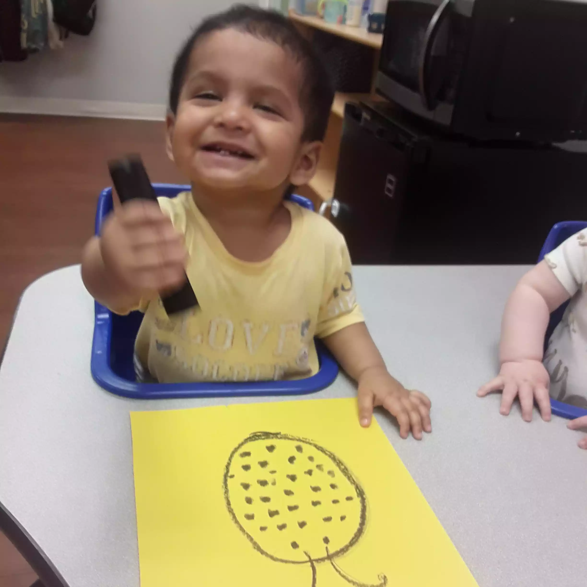 Infant Showing Off Ladybug Art Project - Pinebrook- Preschool - Pinebrook- Preschool Happy toddler sitting in an infant program table seat doing a yellow paper craft activity