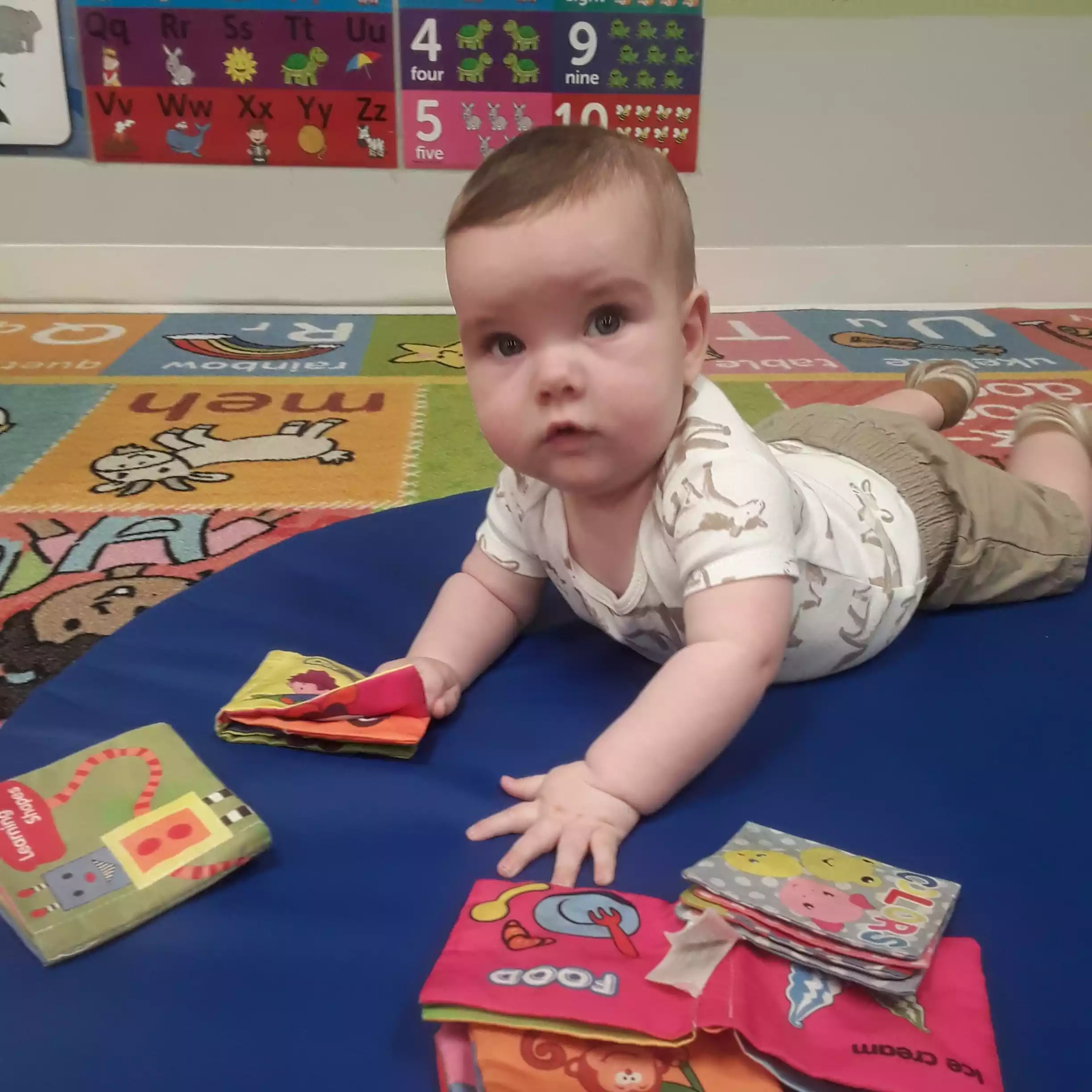 Infant Tummy Time Exploration with Soft Fabric Books - Pinebrook- Preschool - Pinebrook- Preschool Infant enjoying tummy time play on a mat with colorful soft books in classroom