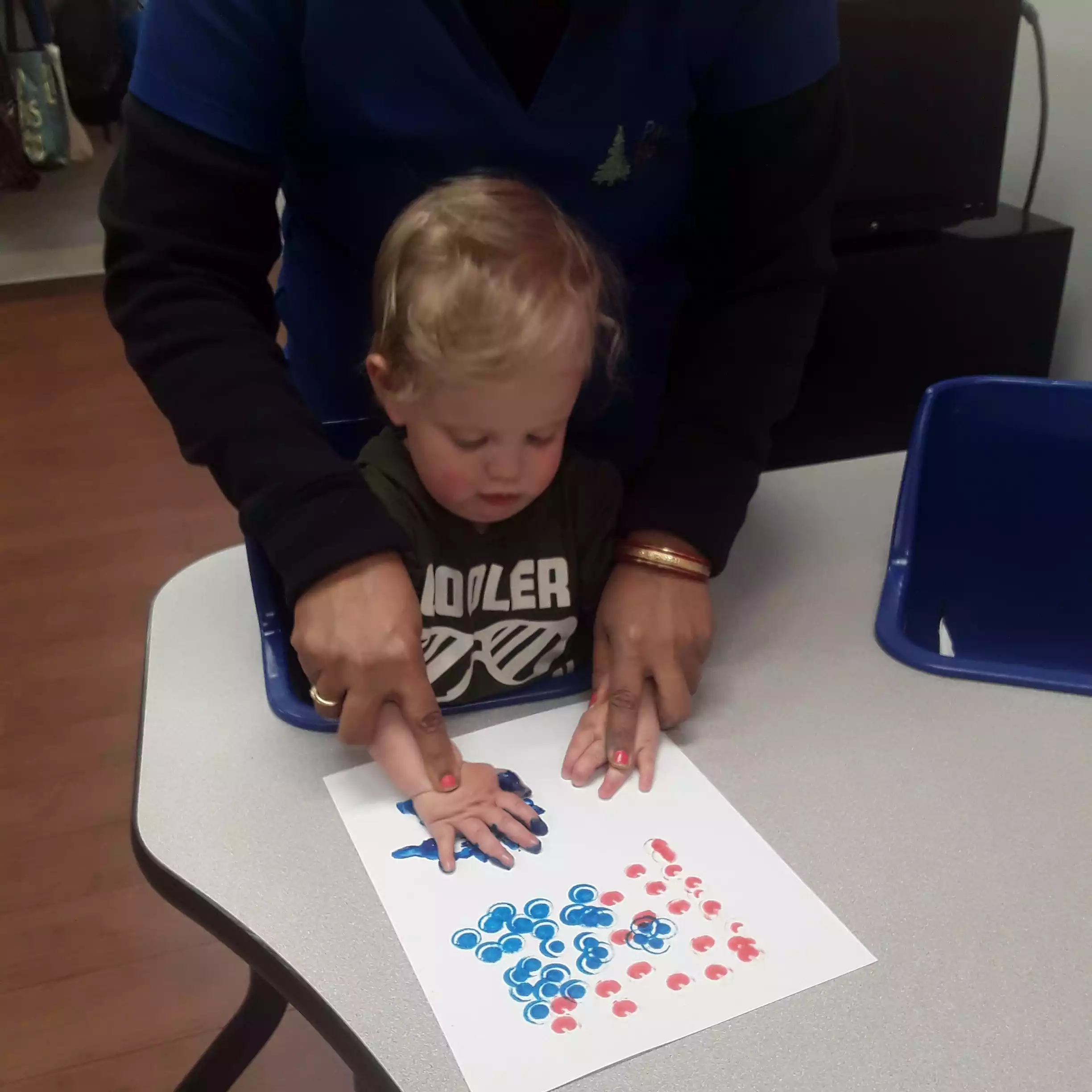 Infant Creating Handprint Painting with Teacher Assistance - Pinebrook- Preschool - Pinebrook- Preschool Teacher helping infant with handprint sensory art project during daycare program