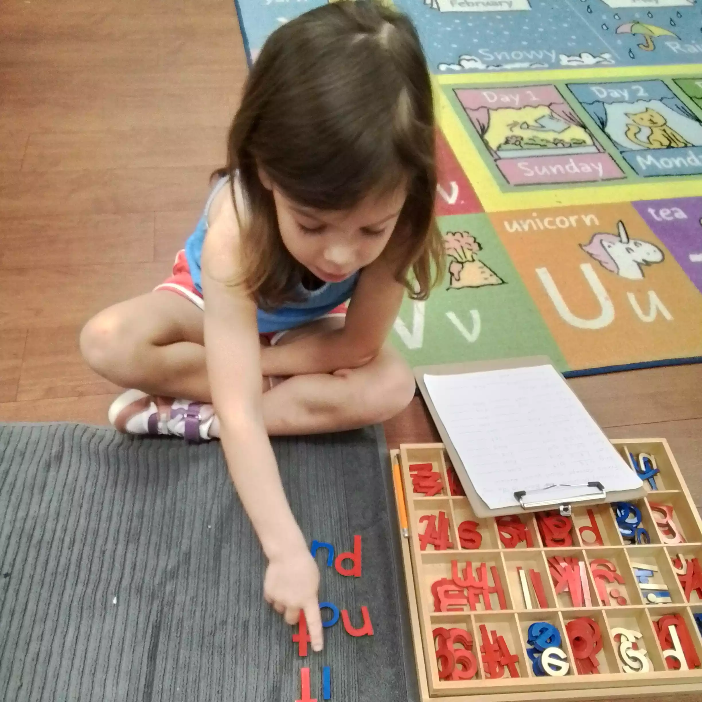 Pinebrook Pre-K Student Practicing Phonics on Reading Mat - Pinebrook- Preschool - Pinebrook- Preschool High-angle view of a young girl pointing at wooden letters spelled out on a mat.