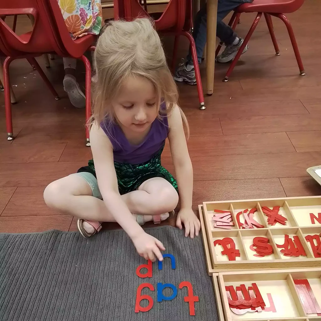 Front view of a young girl sitting cross-legged and pointing to spelled words made of wooden letters.
