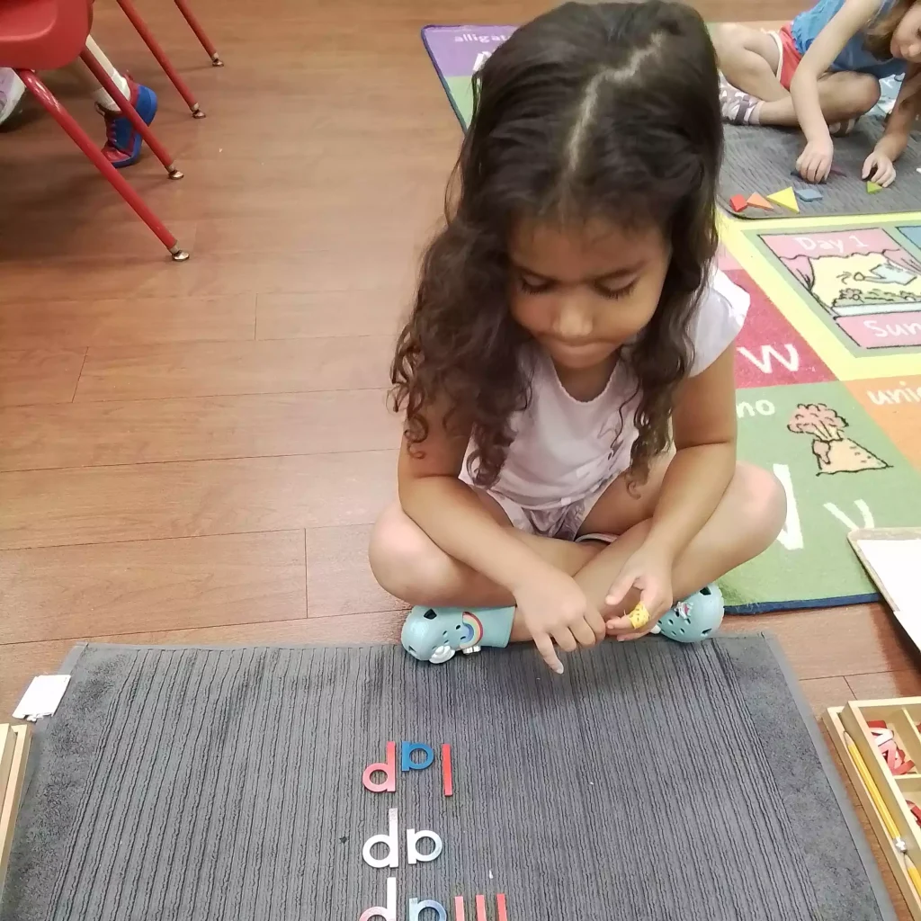 A young girl sitting cross-legged on the floor looking at wooden letters spelling words on a gray mat.