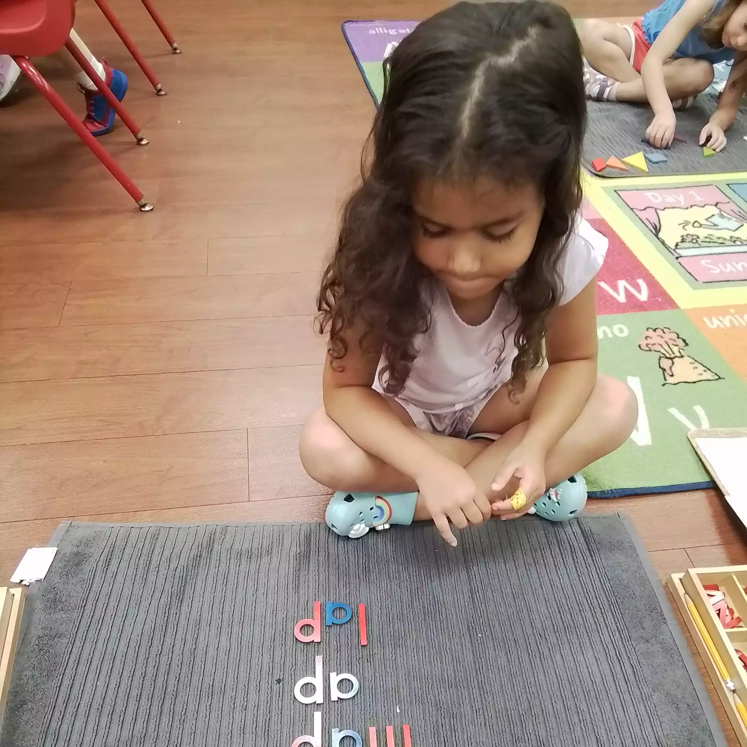 Pinebrook Pre-K Student Studying Wooden Letter Words - Pinebrook- Preschool - Pinebrook- Preschool A young girl sitting cross-legged on the floor looking at wooden letters spelling words on a gray mat.