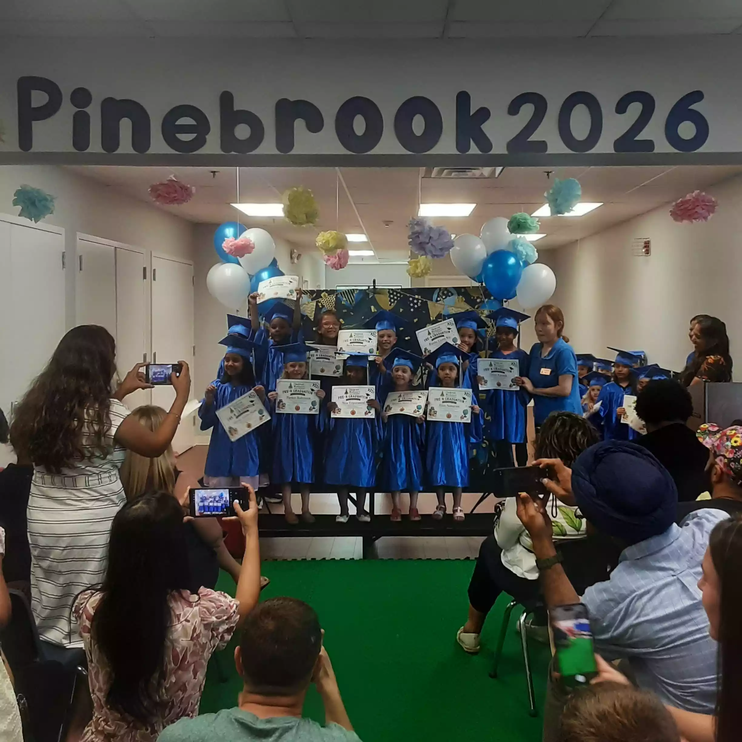 Pinebrook 2026 Pre-K Class Photo on Stage - Pinebrook- Preschool - Pinebrook- Preschool Pre-K graduates in blue caps and gowns holding diplomas on stage
