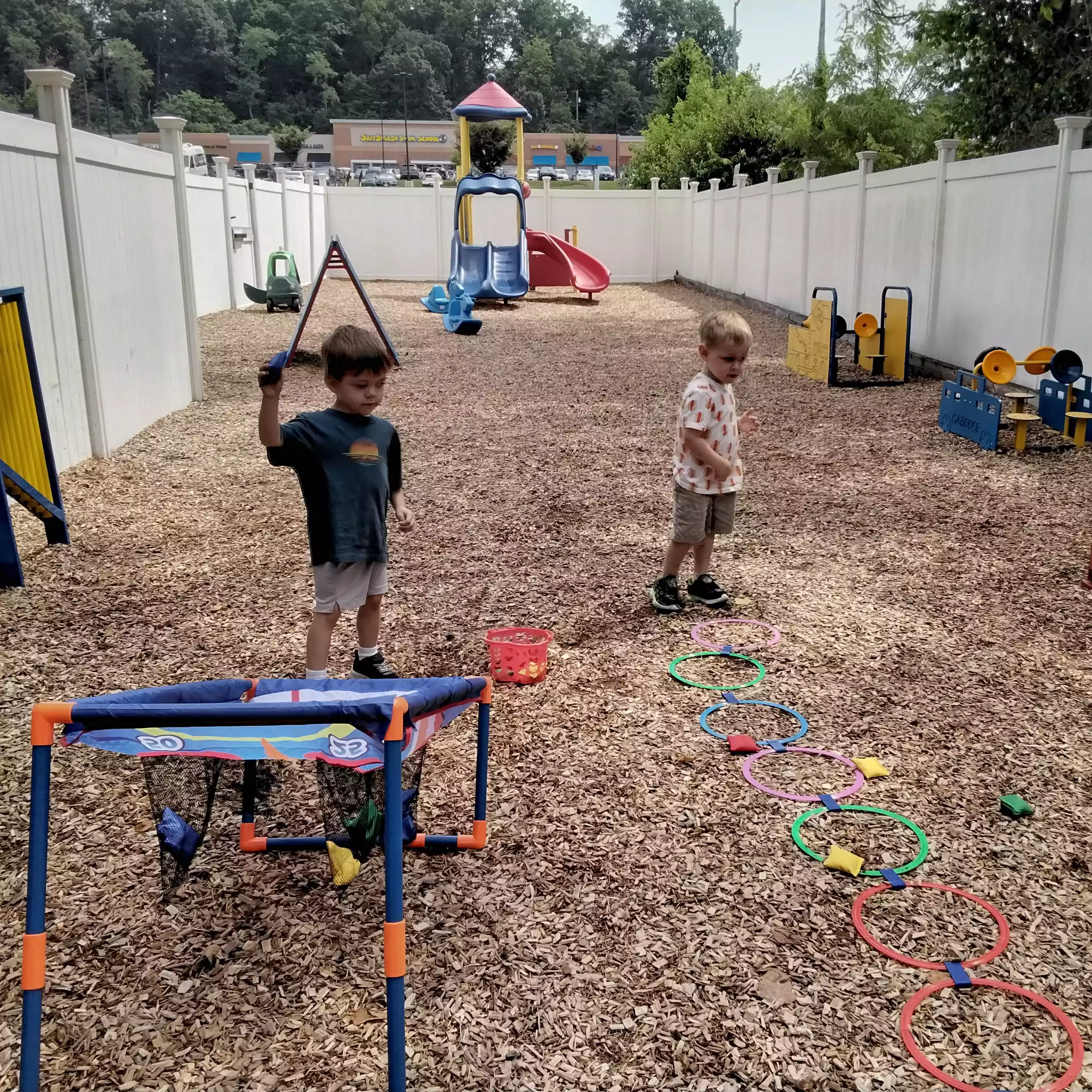 Pinebrook Pre-K Students Playing Playground Beanbag Game - Pinebrook- Preschool - Pinebrook- Preschool Two boys taking turns throwing beanbags at a target on a woodchip playground.