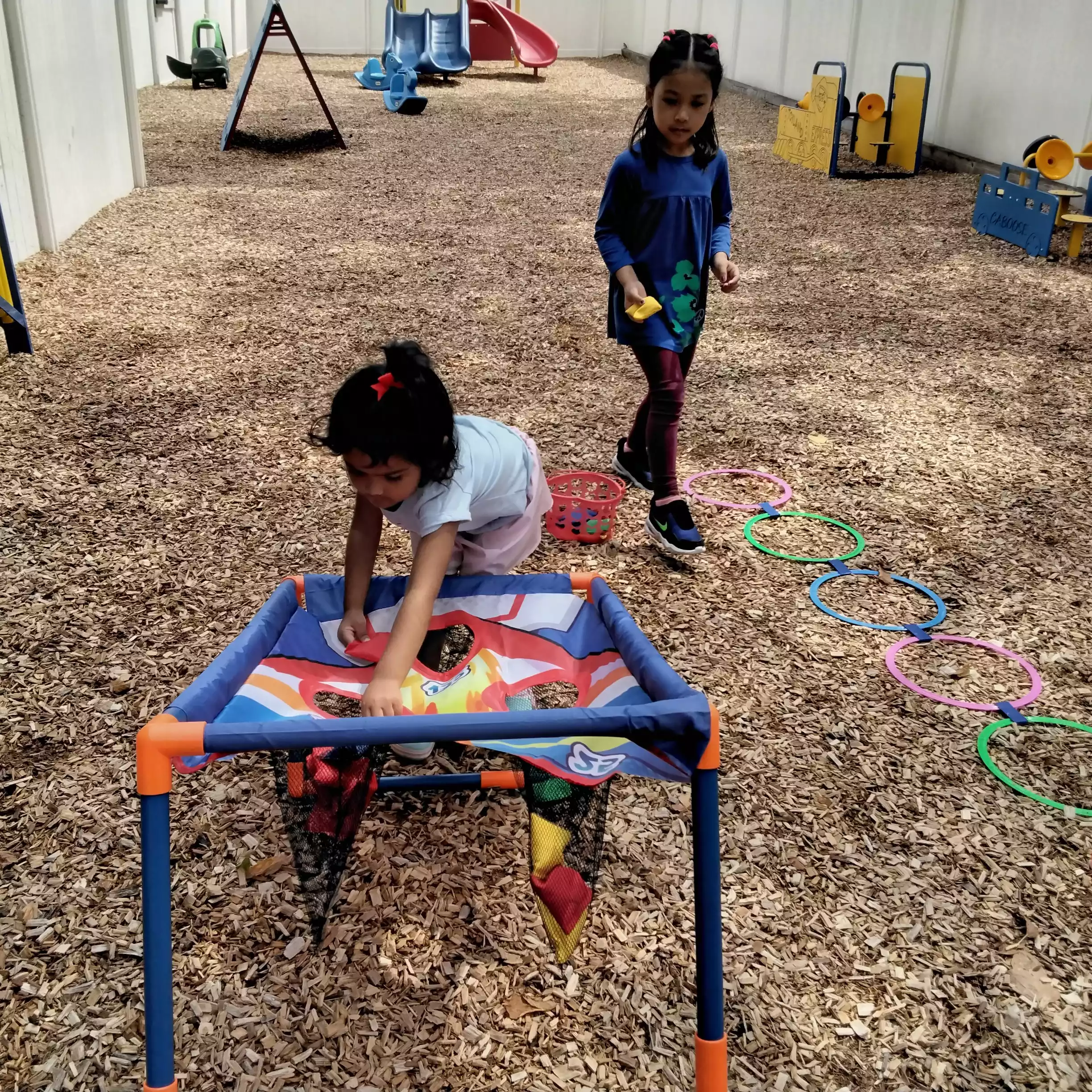 Pinebrook Pre-K Student Reaching into Target Net - Pinebrook- Preschool - Pinebrook- Preschool A young girl reaching into a net target during an outdoor lawn game while another girl walks nearby.