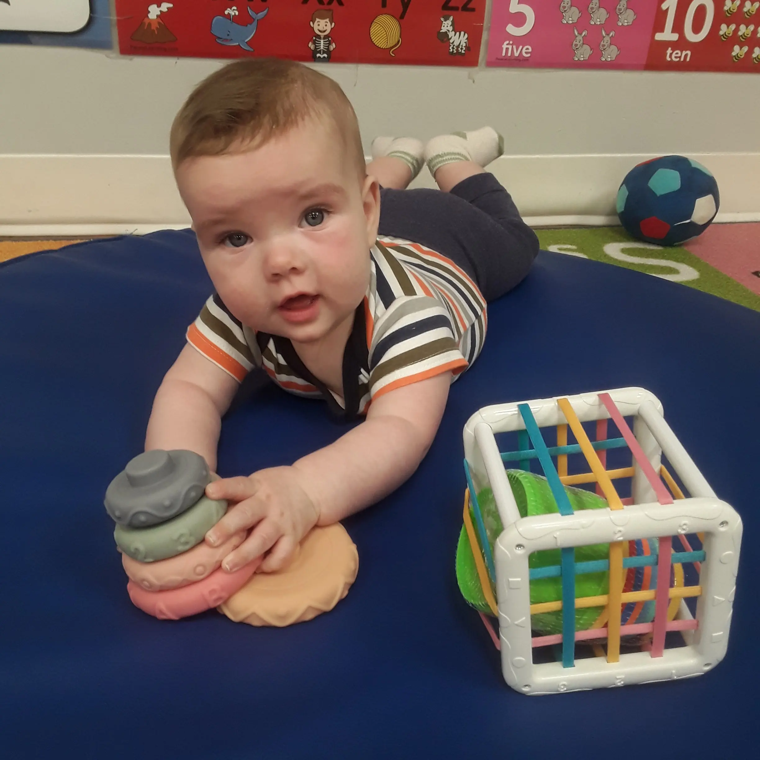 20260515_091727 - Pinebrook Montessori Daycare and Preschool Baby lying on a blue mat, gripping a soft gray toy in one hand with a colorful activity cube nearby, classroom posters visible in the background.