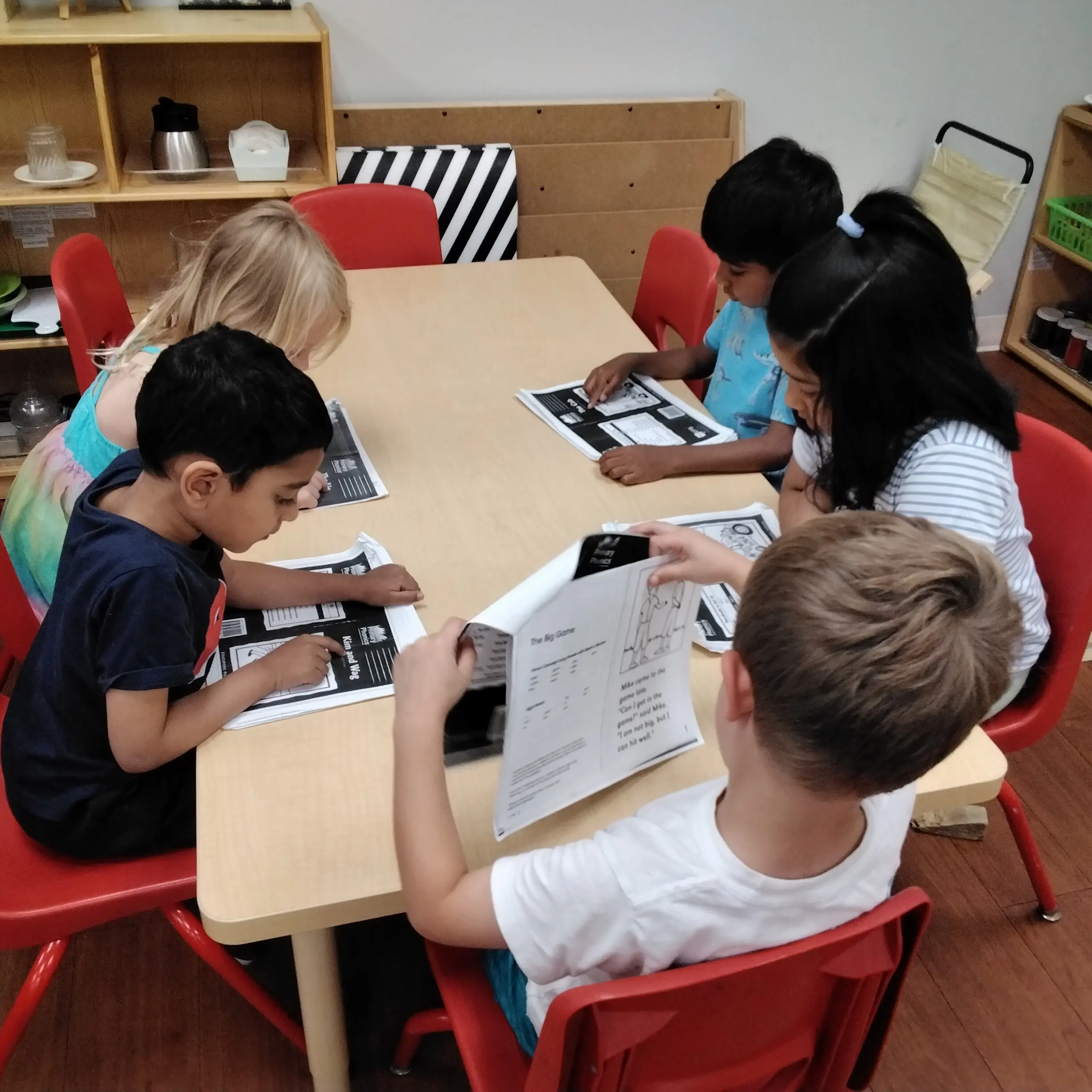 Children seated around a light wood table, reading worksheets in a classroom with red chairs nearby