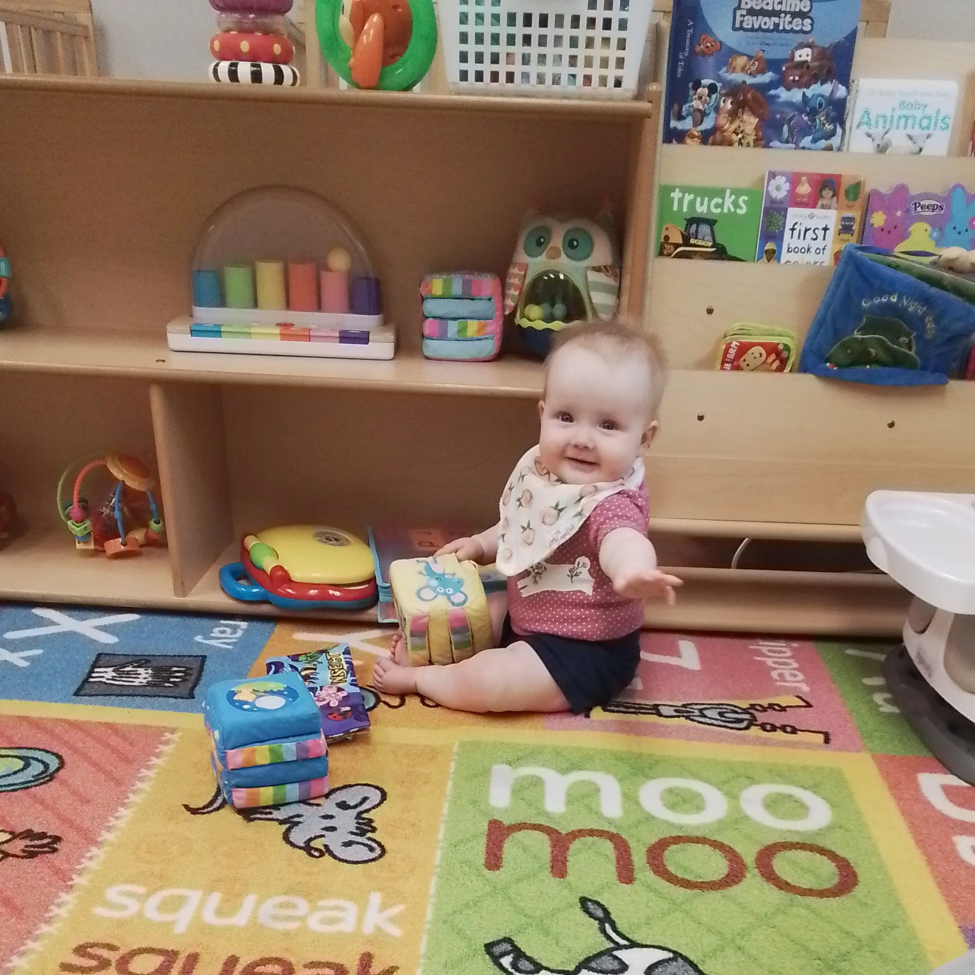 20260623_145428 - Pinebrook Montessori Daycare and Preschool Smiling baby sits on a colorful rug with soft blocks, shelves and children's books visible in a playroom background.