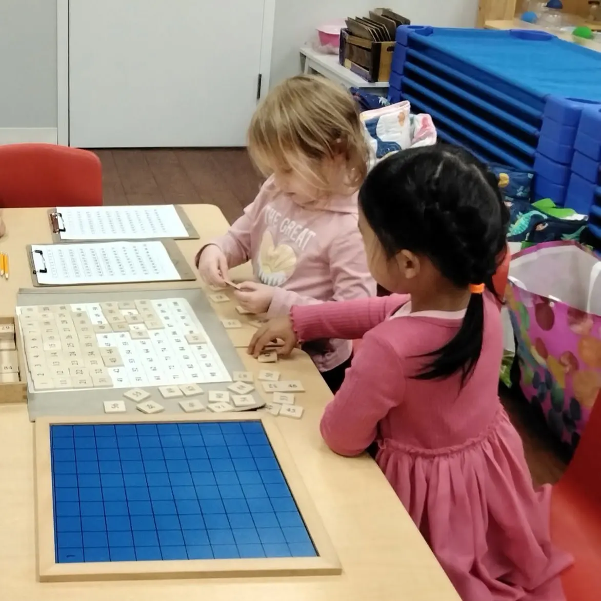 646765181 - Pinebrook Montessori Daycare and Preschool - Pinebrook Montessori Daycare and Preschool Two young girls in pink sit at a table building words with wooden letter tiles and a blue grid mat nearby in a classroom.