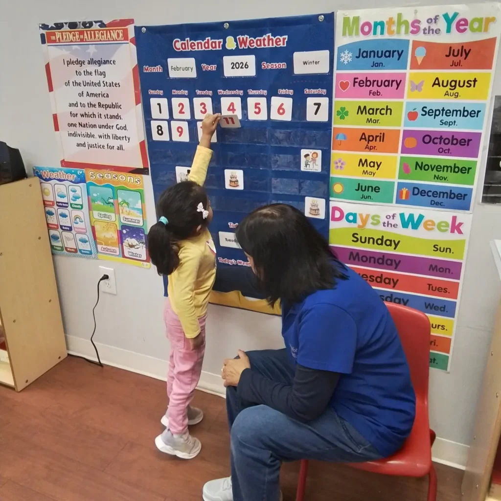 Young girl places a number card on a large blue 'Calendar & Weather' board as a teacher watches in a classroom moment.
