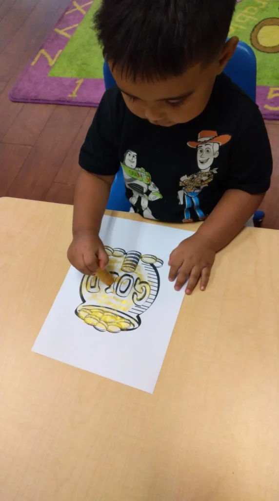 Child sits at a table coloring a jar of coins on a paper with a yellow crayon, wearing a black t-shirt with Toy Story characters.