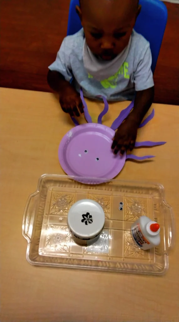 Young child at a table making a purple spider craft using a paper plate and purple paper strips as legs with googly eyes.
