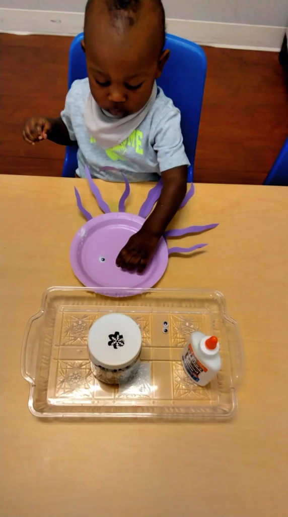 Young child sits at a table gluing a purple paper sun craft with wavy rays. nearby glue bottle and supplies visible.