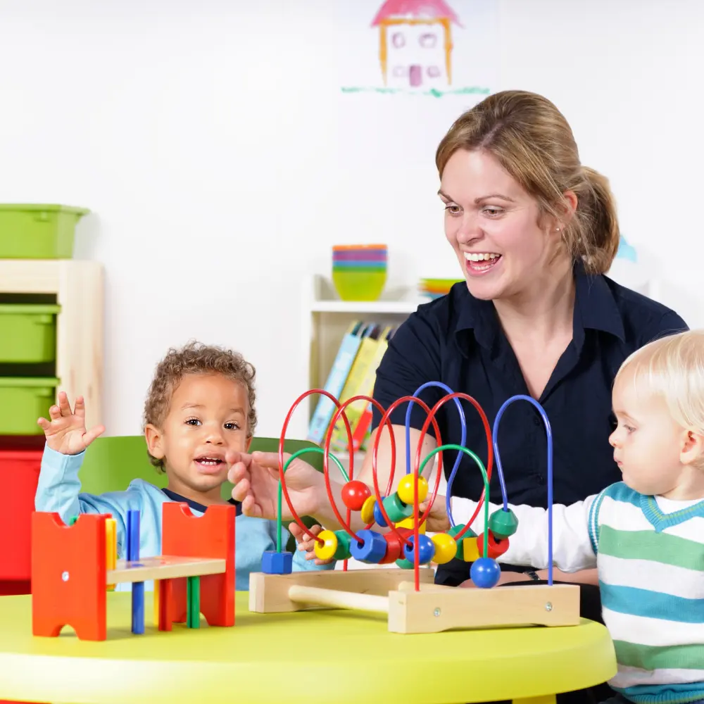 Alleviating Frustration through Speech Teacher helps two toddlers as they play with a bead maze and colorful blocks at a round yellow table in a bright classroom.