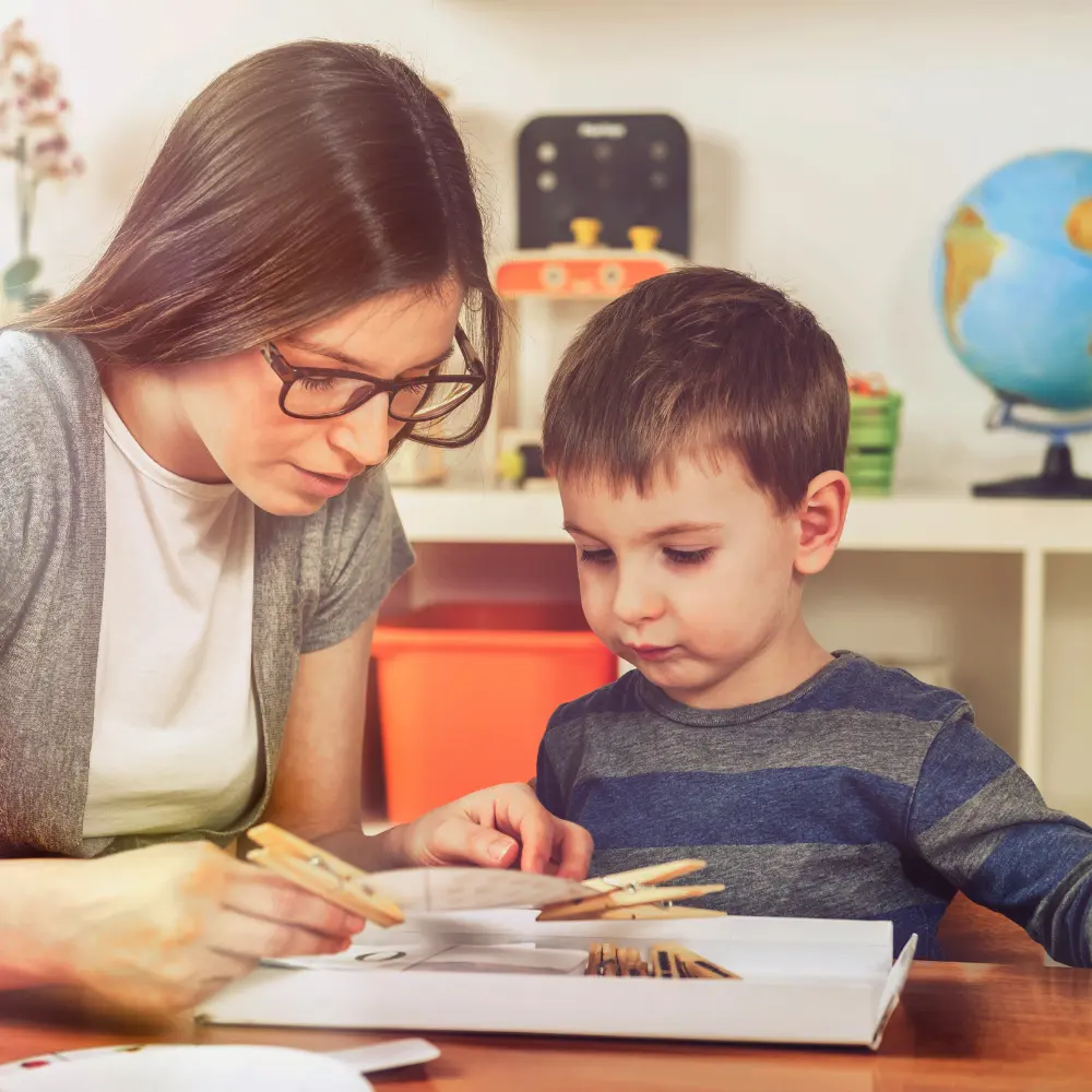 Authentic Trust & Parental Involvement Woman helps a child with a craft project, clipping wooden clothespins onto a page at a table in a classroom-like setting.