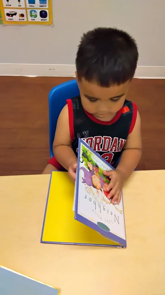 Young child wearing a red and black sleeveless shirt sits at a yellow table, flipping through a colorful picture book while looking down at the pages on a blue chair nearby.