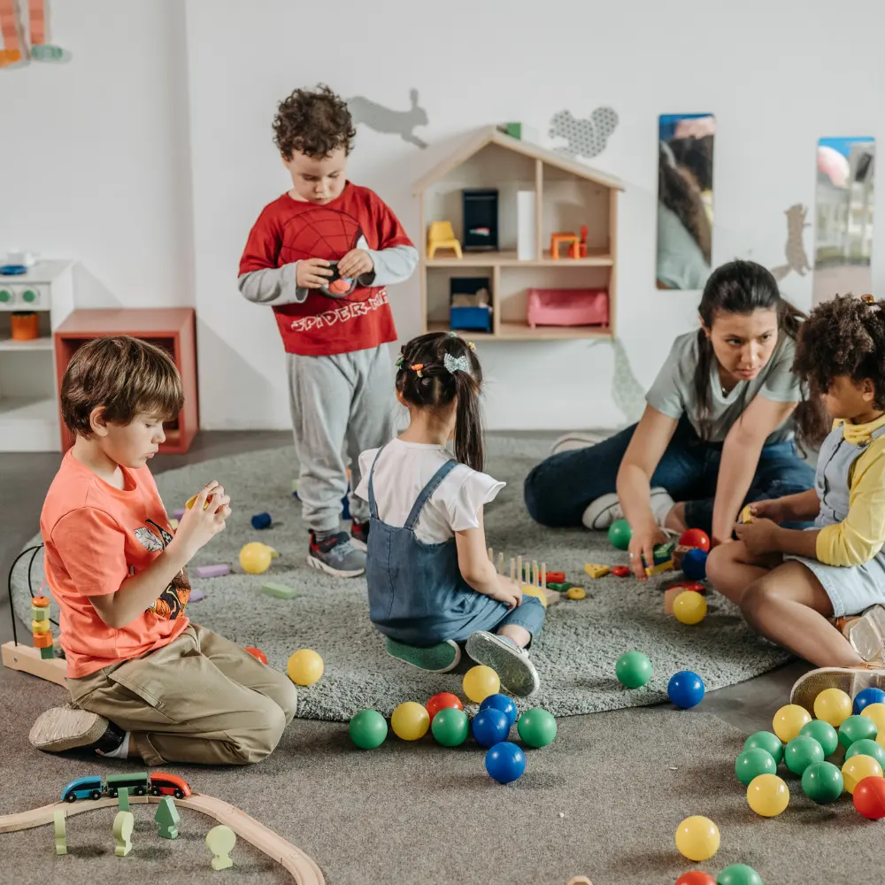 Teacher kneels on a carpeted playroom floor, guiding kids as they play with colorful balls and wooden toys on a rug.