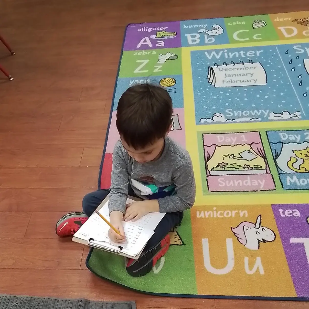 A young child sits on a colorful alphabet-themed classroom rug and writes in a notebook with a pencil.