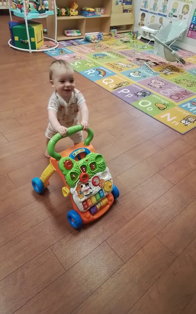 Smiling toddler pushing a colorful activity walker on a wooden floor in a bright playroom.