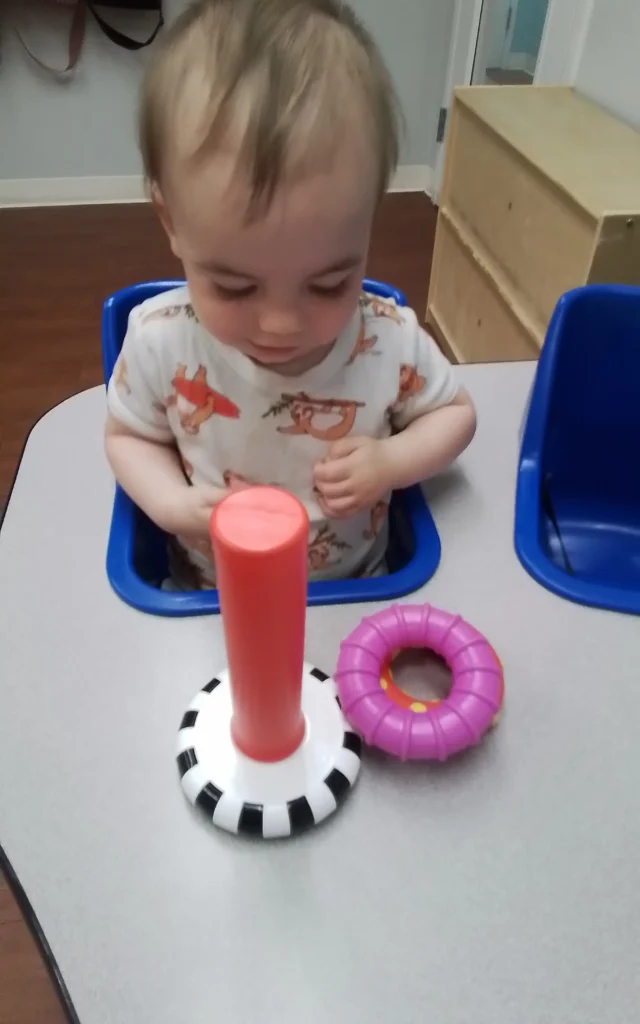 Toddler in a blue high chair at a table, playing with an orange cylinder and a pink ring toy on the table surface.