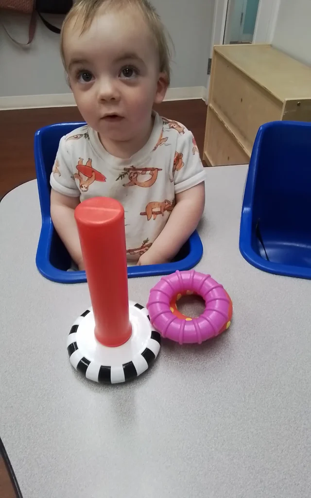 Toddler in a white animal-print shirt sits in a blue chair at a table, looking toward the camera, with a red cylindrical toy and a purple ring-shaped toy in front.