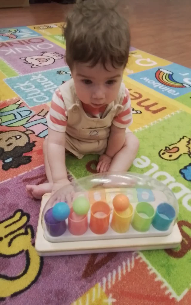 Young child sitting on a colorful alphabet rug, reaching toward a clear toy with multicolored cups and balls on the floor.