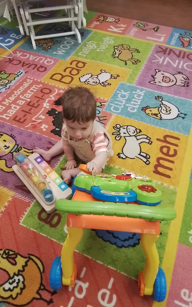 Young child sitting on a bright alphabet rug, playing with a small white tablet-style toy and a colorful pushing toy truck nearby.