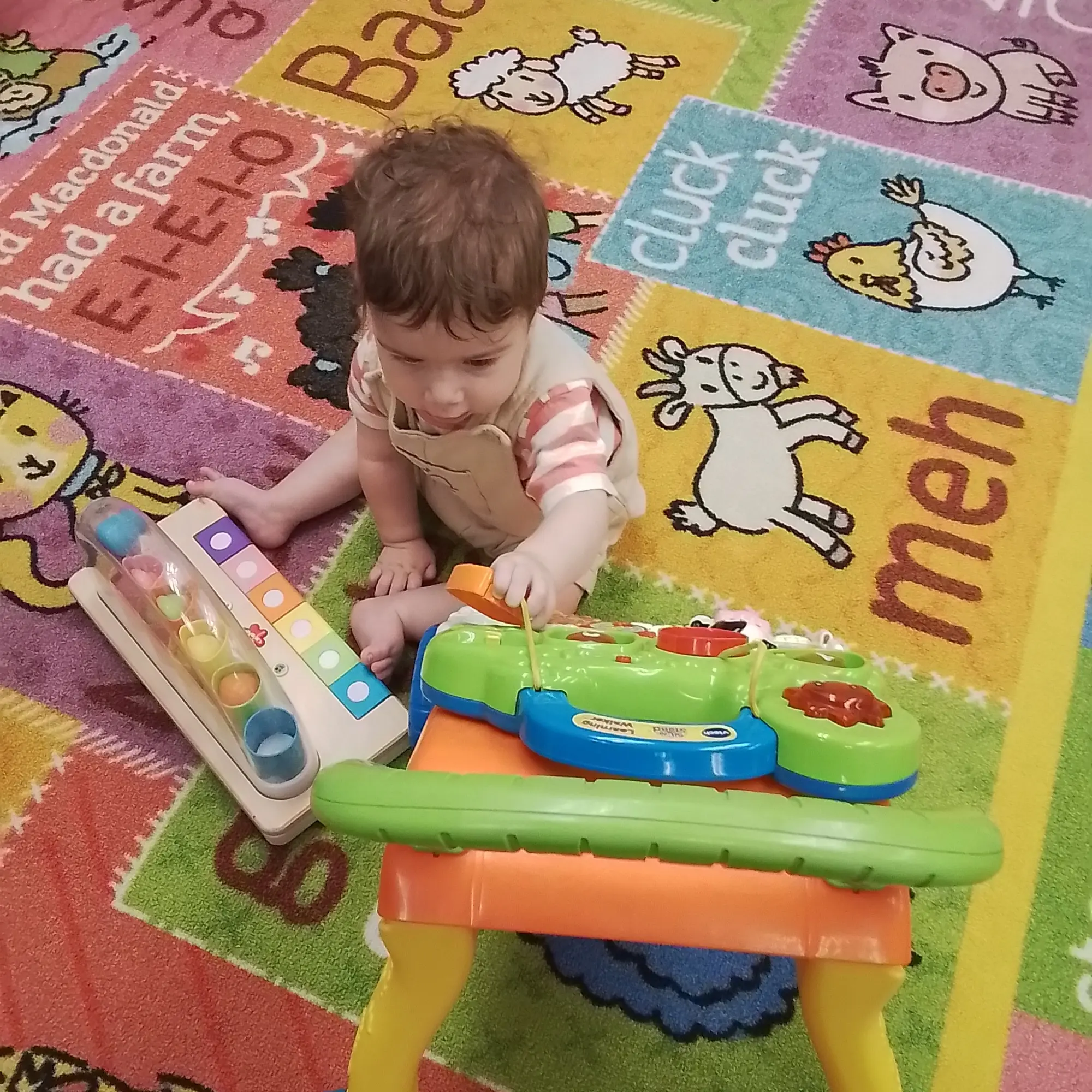 Infant-20260805_084726 - Pinebrook Montessori Daycare and Preschool Young child sits on a colorful cartoon rug, playing with educational toy blocks and a shape/color matching toy.