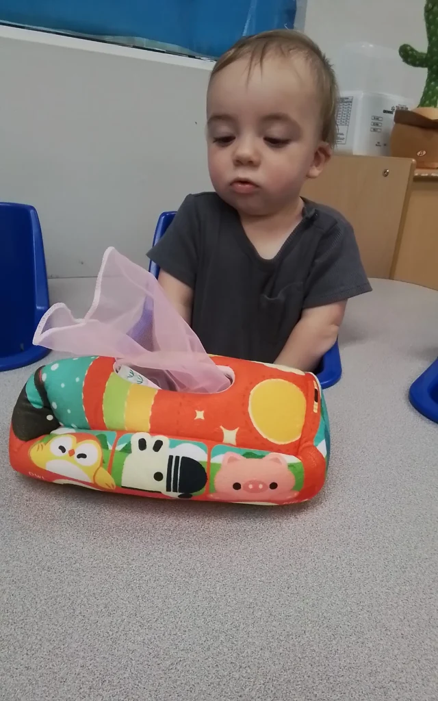 Toddler with short hair sits in a blue chair, wearing a dark gray shirt, looking down at a colorful inflatable neck pillow with animal prints and a pink tissue sticking out.