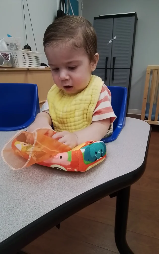 Baby seated in a blue high chair at a gray table, wearing a yellow bib and playing with a colorful inflatable toy.