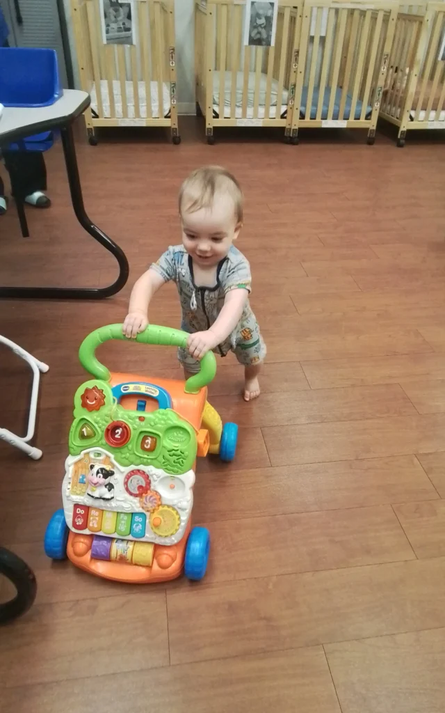Young child pushing a bright plastic activity walker with buttons and animal pictures in a playroom with wooden cribs in the background.