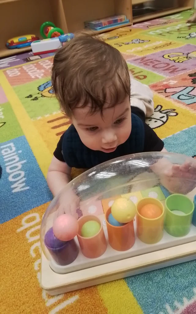 Baby sits on colorful rug, reaching into a clear bubble toy filled with pastel balls and small cups nearby for play.