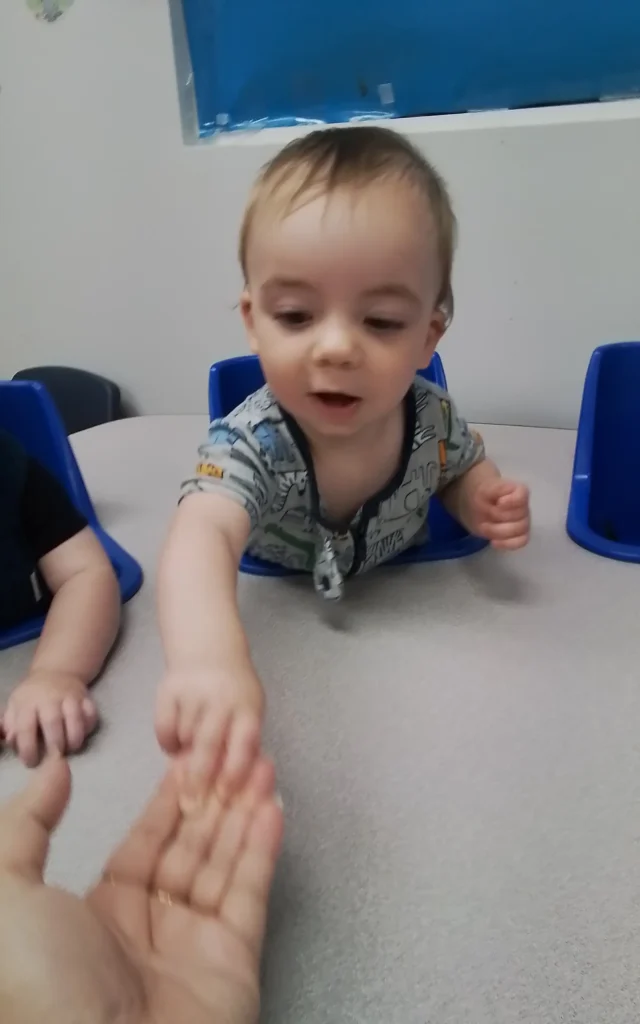 Toddler reaching forward with an outstretched hand at a table, blue chairs in the background.