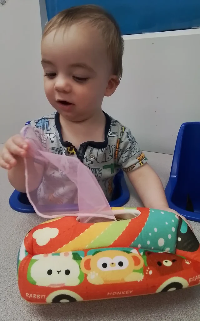 Toddler sitting in a blue high chair, handling a pink feeding bib beside a colorful animal-themed cushion.