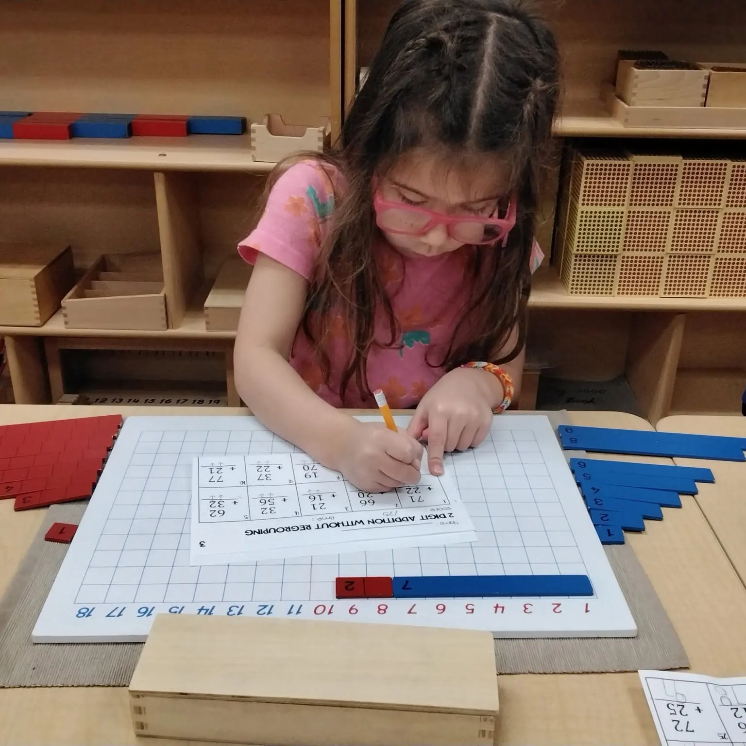 Young girl in a pink shirt and glasses writes with a pencil on a math worksheet at a desk, surrounded by base-10 blocks and grids for counting.