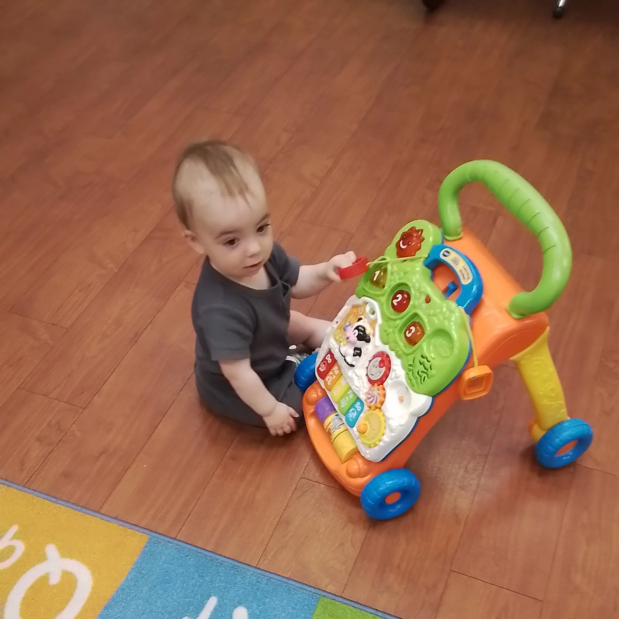 Young child stands at a bright, multi‑colored activity center on a wooden floor, touching the toy components.