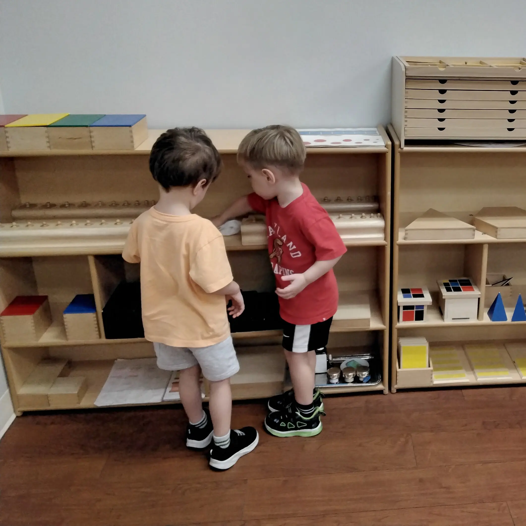 (Practical Life - Cleaning) - Pinebrook Montessori Daycare and Preschool Three children stand at a wooden shelf in a classroom, looking at or playing with block-like educational materials.