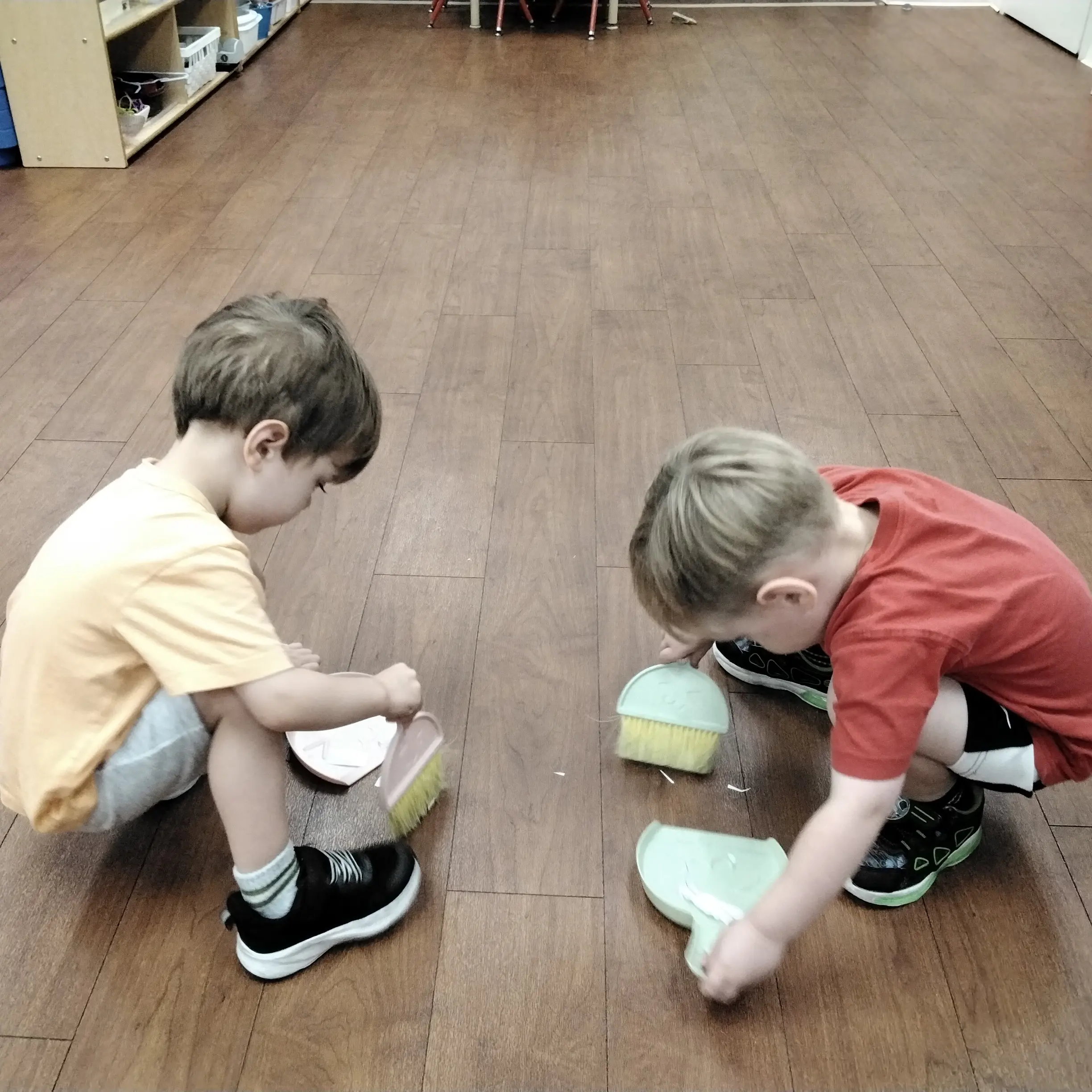 Two children sit on a wood floor, painting plastic cups with brushes in a classroom setting.