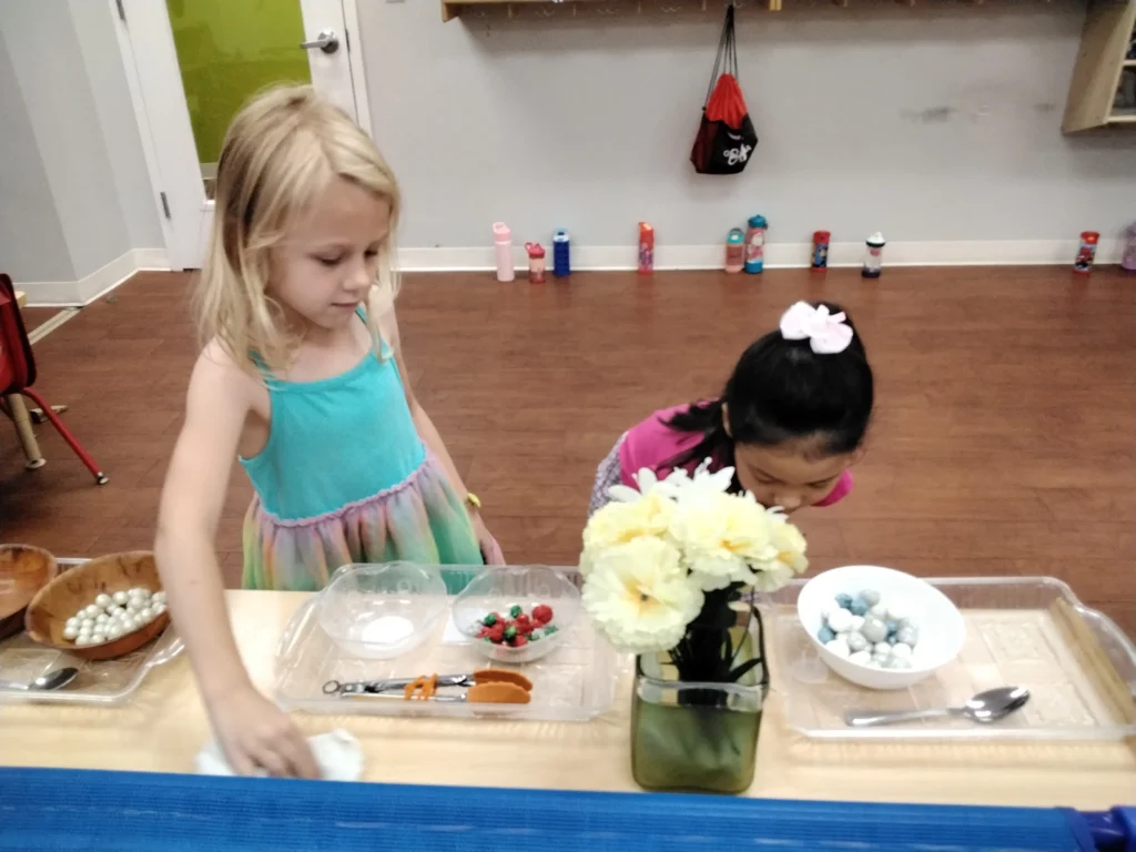 Two young girls work at a craft table with bead trays, scissors, and flowers in a classroom setting.