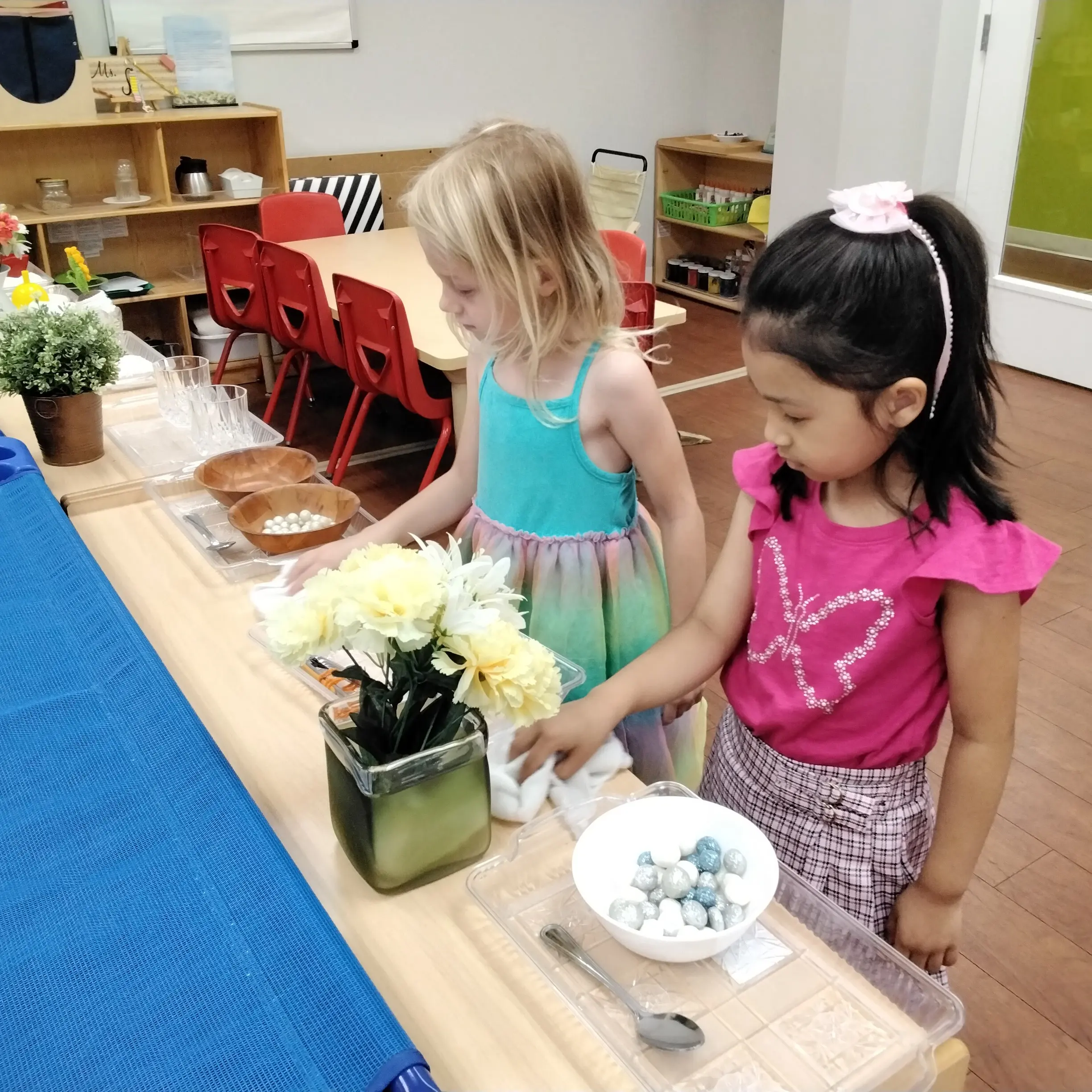 Two young girls in colorful dresses stand at a table with bowls of decorative items and flowers in a classroom setting.
