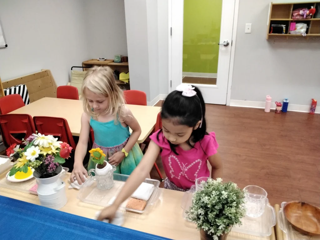 Two young girls stand at a table in a classroom, crafting with flowers and small white vases nearby.