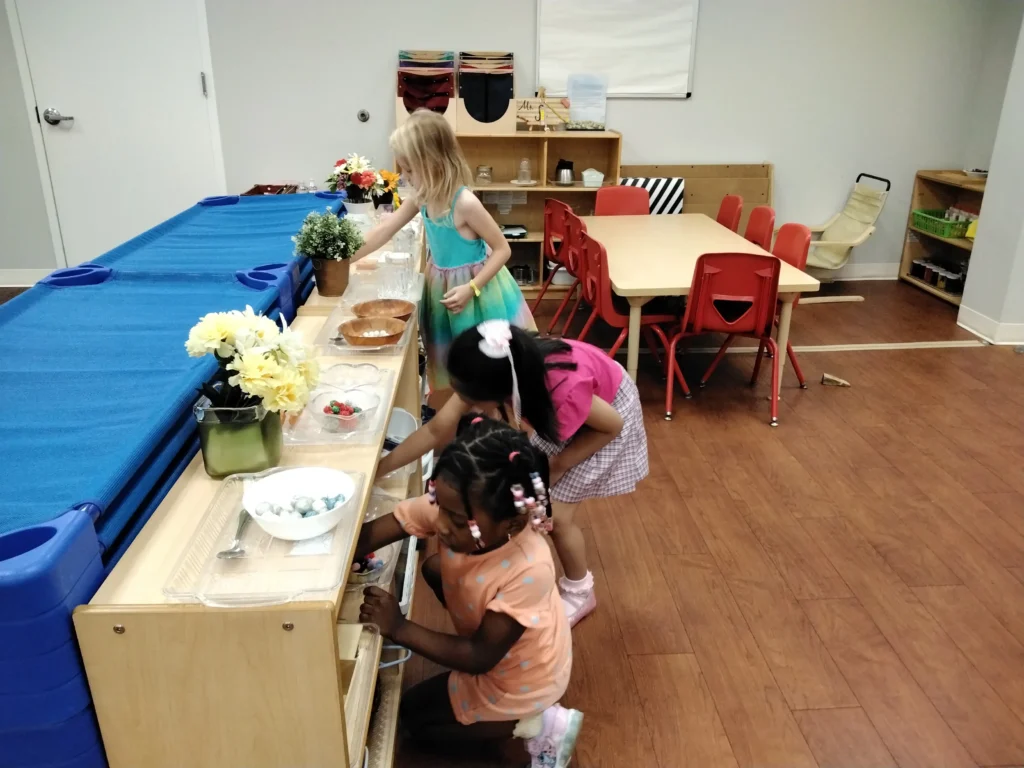 Children at a dessert bar in a classroom, selecting treats from clear containers along a wooden counter with flowers nearby