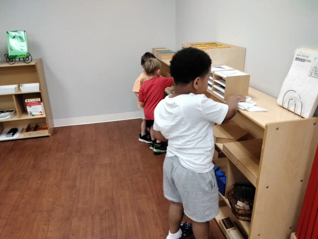 Three children stand at a wooden classroom counter, facing storage cubbies and papers in a light gray room.