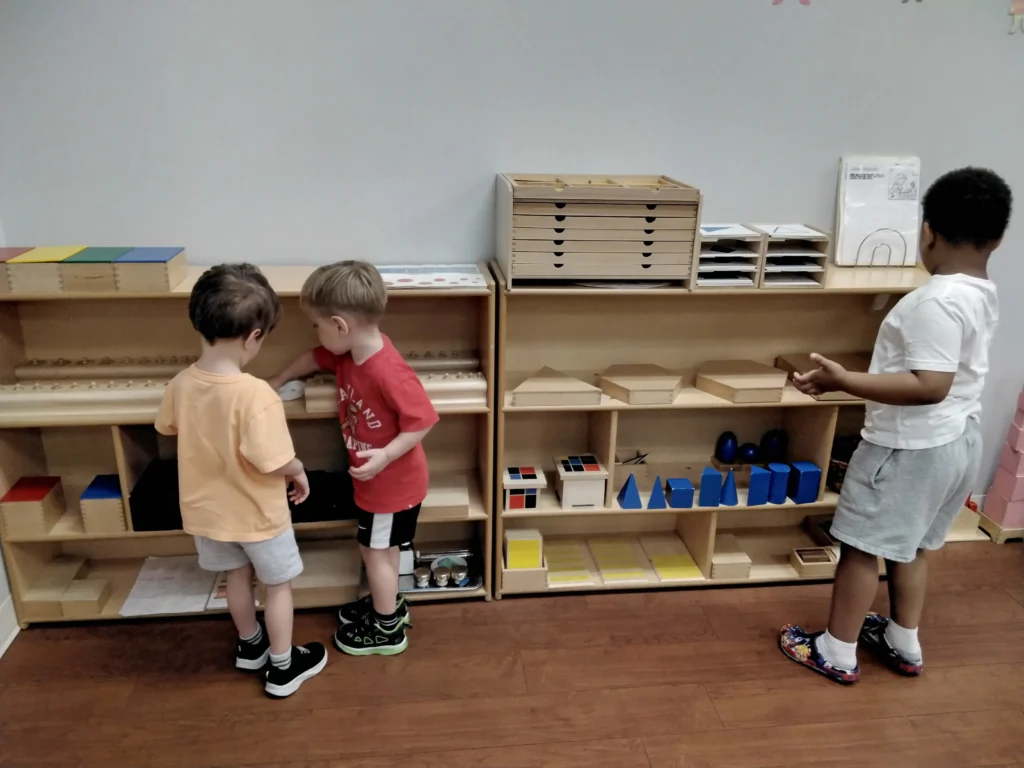 Three children and a supervising adult stand by wooden shelves in a classroom, playing with geometric blocks and sorting trays on the shelves.