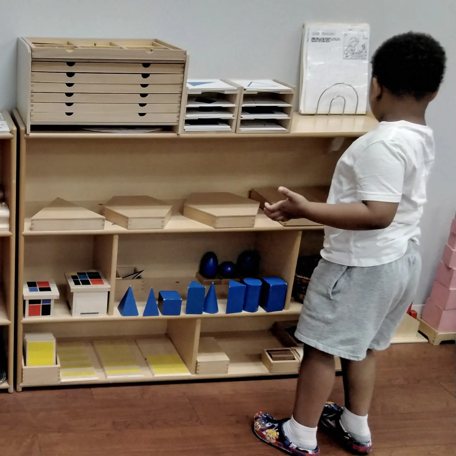 Young boy in white shirt and gray shorts stands at a shelf of wooden educational blocks and shape manipulatives in a classroom.