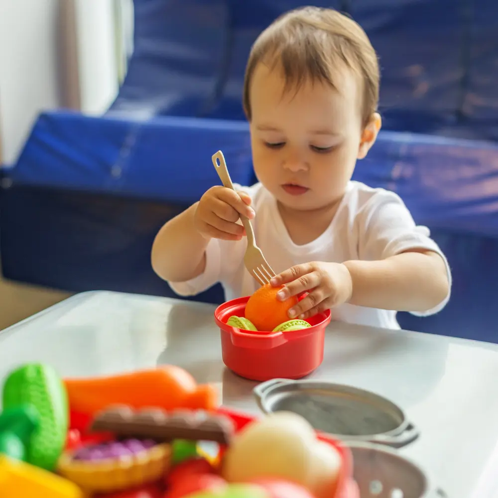 Practical Life & Self-Care - Pinebrook Montessori Daycare and Preschool Child in a white shirt sits at a desk reading a colorful picture book on blue chair in a classroom setting