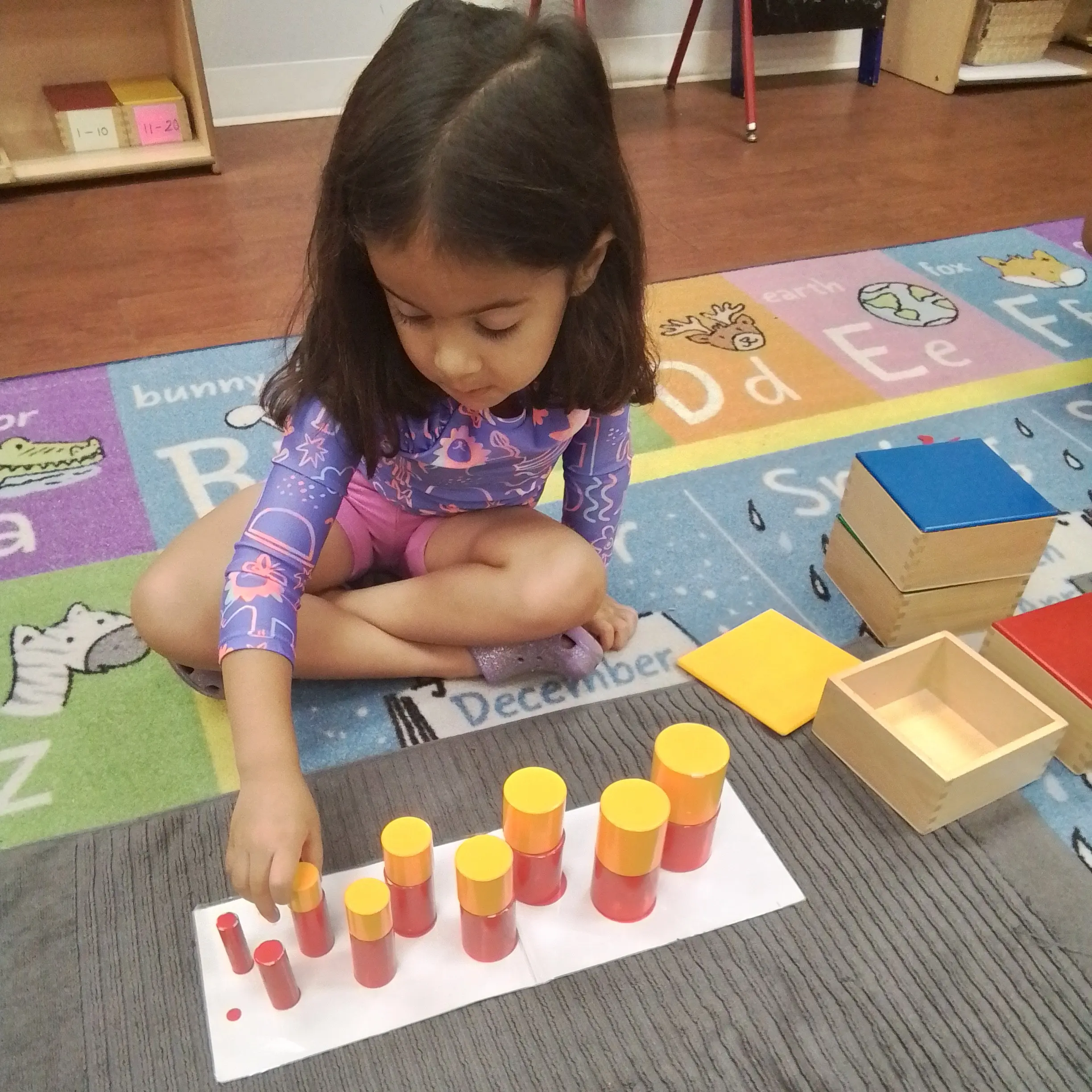 Young child stacking orange and yellow cylindrical blocks on a white board during a classroom activity.