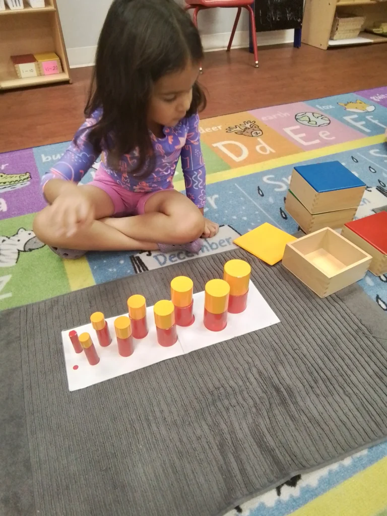 Young girl sits cross-legged on a colorful classroom rug, arranging red and yellow stacking cylinders on a white mat.