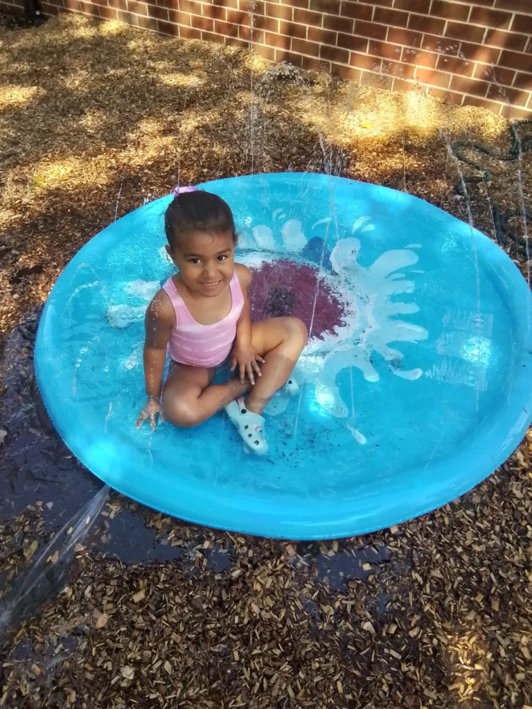Young girl in a pink swimsuit sits in a small blue plastic pool with water spraying around her in a sunny yard with wood chips and a brick wall behind.